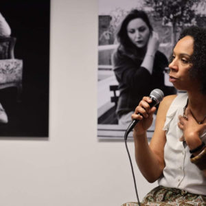photo de Nafi Yao micro à la main lors de présentation du livre au Centre Culturel de Herstal en Belgique. En arrière plan, une photo de Julie Foulon, Girleek.