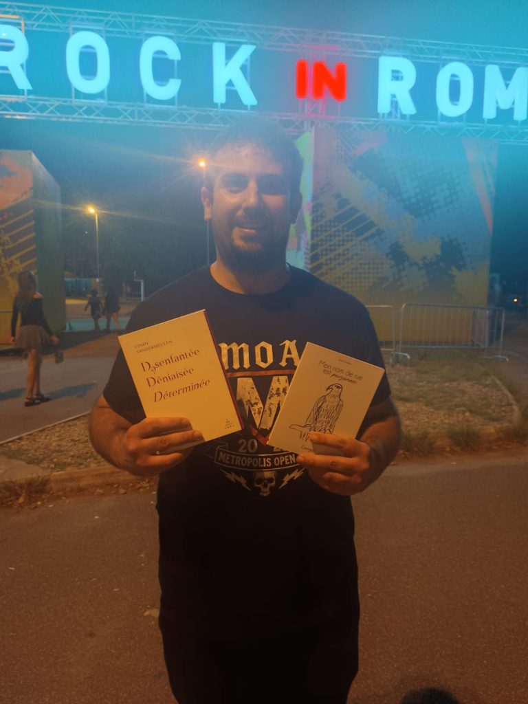 Photo d'un lecteur devant le festival rock in roma avec en main le livre de Tessy Oudèna, mon nom de rue est personne paru chez courgette editions et le livre Désenfantée, Déterminée, Déniaisée de Cindy Vandermeulen paru chez Abrapalabra Editions.