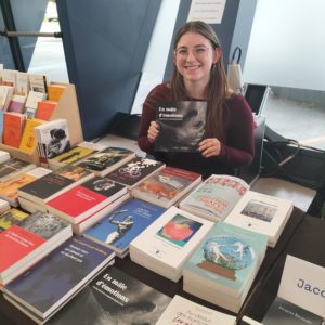 La stagiaire de Courgette Éditions sourit derrière un stand de livres, tenant fièrement l’ouvrage En mâle d’émotions. Autour d’elle, une table généreusement couverte de titres variés crée une atmosphère conviviale et passionnée, typique des salons du livre où l’édition indépendante rayonne.