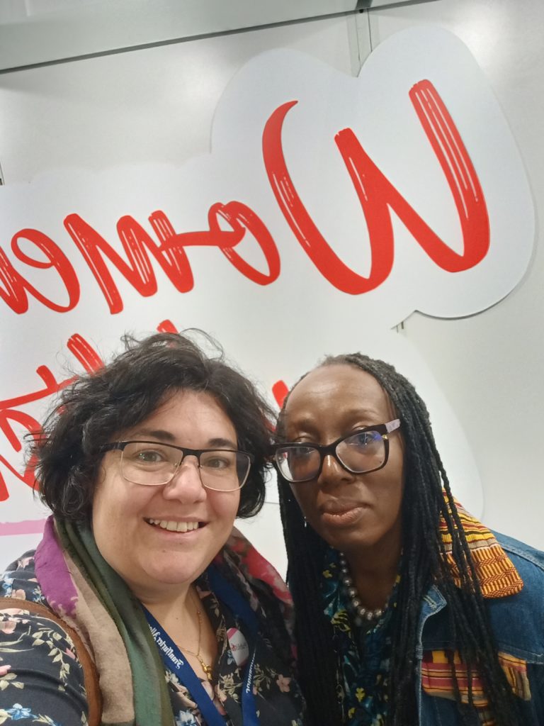 Deux femmes souriantes, Cindy Vandermeulen à gauche et Ama Dadson à droite, posent pour un selfie devant une grande enseigne rouge stylisée portant le mot "Women", lors d’un événement dédié à l’empowerment féminin. Elles portent des badges et des tenues colorées, suggérant une ambiance conviviale et professionnelle.