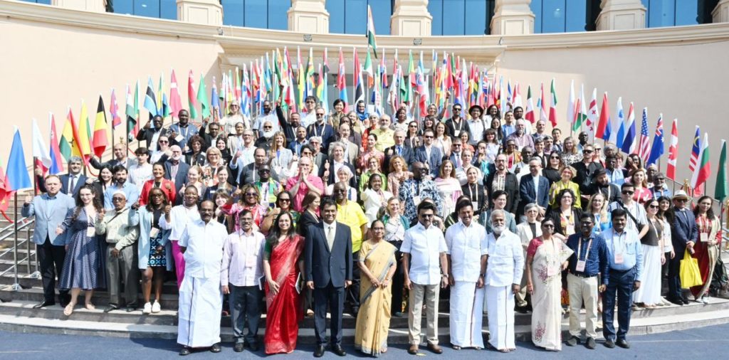 Photo de groupe prise à Chennai, en Inde, lors de la Chennai International Book Fair (CIBF) : plus d’une centaine de représentants et représentantes de pays du monde entier posent ensemble devant un bâtiment officiel, entourés de nombreux drapeaux nationaux. Les personnes sont disposées sur plusieurs rangées, souriantes, dans une grande diversité de tenues traditionnelles et contemporaines. En bas à droite, au deuxième rang, cindy vandermeulen porte une robe noire, jaune et rouge