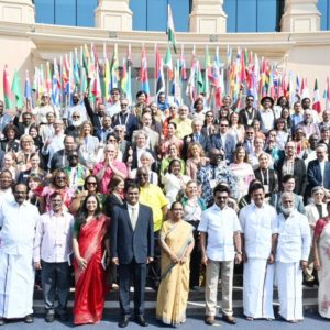 Photo de groupe prise à Chennai, en Inde, lors de la Chennai International Book Fair (CIBF) : plus d’une centaine de représentants et représentantes de pays du monde entier posent ensemble devant un bâtiment officiel, entourés de nombreux drapeaux nationaux. Les personnes sont disposées sur plusieurs rangées, souriantes, dans une grande diversité de tenues traditionnelles et contemporaines. En bas à droite, au deuxième rang, cindy vandermeulen porte une robe noire, jaune et rouge