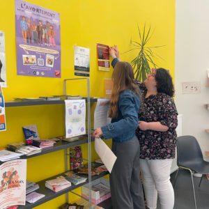 photo prise au centre de planning familial de herstal où l'on voit philippine (stagiaire) et cindy vandermeulen (éditrice) en train de coller une affiche pour la sortie du livre présomption de culpabilité