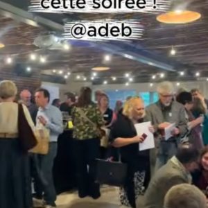 capture d'écran d'une vidéo prise lors de l'assemblée générale de l'adeb on l'on voit plusieurs personnes discuter et porter des toasts.