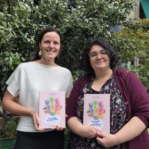 photo de face de barbara de munnynck avec le livre un monde de merveilles édité chez courgette éditions en main et à droite, cindy vandermeulen, éditrice de courgette éditions avec en main, le livre de wereld is een wonderkamer édité chez pelckmans uitgevers, la version néerlandophone avec en arrière plan des plantes vertes.