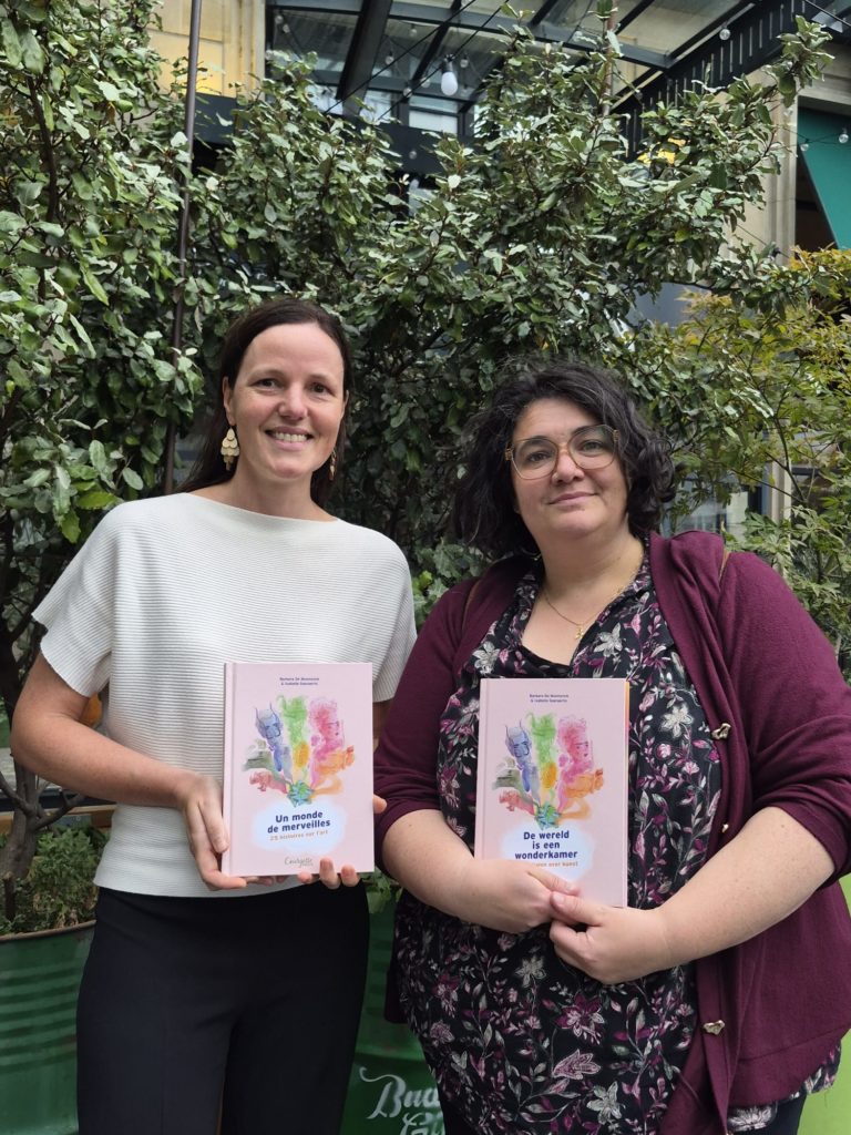 photo de face de barbara de munnynck avec le livre un monde de merveilles édité chez courgette éditions en main et à droite, cindy vandermeulen, éditrice de courgette éditions avec en main, le livre de wereld is een wonderkamer édité chez pelckmans uitgevers, la version néerlandophone avec en arrière plan des plantes vertes.
